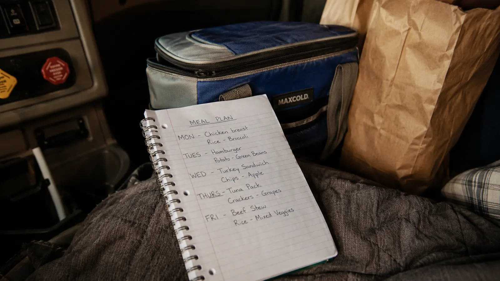 A handwritten weekly meal prep list on a notepad next to a small cooler and a bag of groceries in a semi truck sleeper