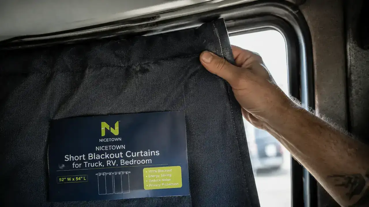 Hand holding NICETOWN blackout curtain panel next to a sleeper cab bunk window, checking the fit against the frame
